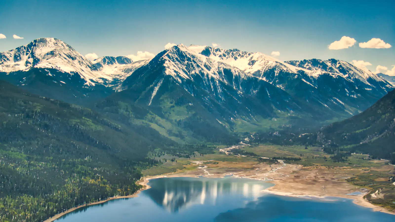 A teal alpine lake and braided valley beneath snow-capped mountains, seen from above.